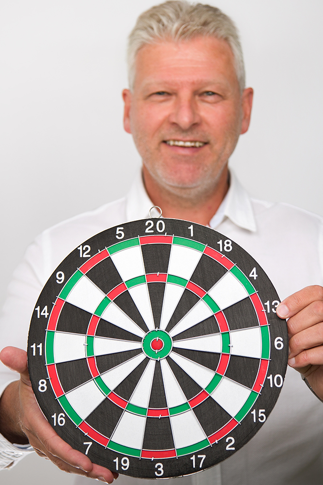 DEBATIN_Produktsparten_Headerbild_DEBADEINS Christophe Houdé is wearing a white shirt and holding up a colorful dartboard.