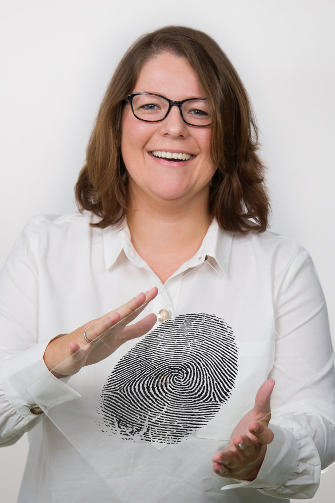 DEBATIN_Produktsparten_Headerbild_DEBADEINS_Stricker_Fingerabdruck Veronika Stricker is wearing a white blouse and holding up a fingerprint engraved into a plexiglass panel.