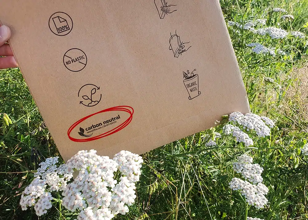 DEBAPOST-Paper-Second-Life-carbon-neutral-Logo-change A brown paper mailing bag (DEBAPOST Paper Second Life) with sustainability logos and labelling is pictured in a meadow, surrounded by white flowers. The logos include “100% paper”, “no plastic", “organic waste”. In addition, there is a “carbon neutral” logo, circled in red.