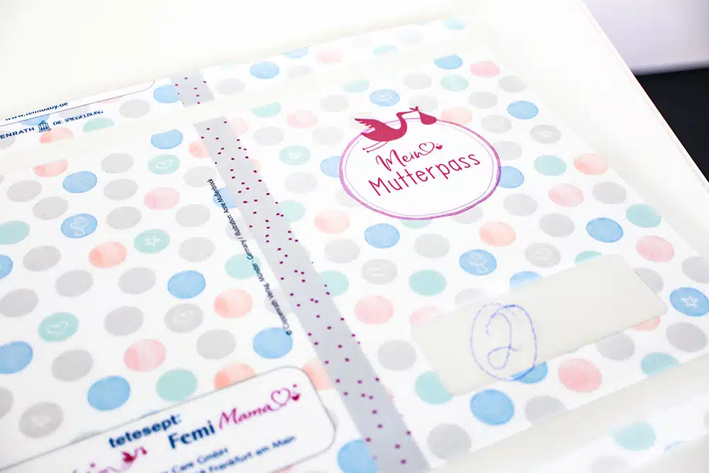 IMG_0101 Close-up of a colourful folder printed using the DEBAPRINT flexographic process by DEBATIN, titled My Maternity Record. The design features a pattern of circles in pink, blue, and grey, along with a round logo depicting a pink stork. The folder protects the maternity record, with personal details partially visible, and displays the "tetesept Femi Mama" logo at the bottom.