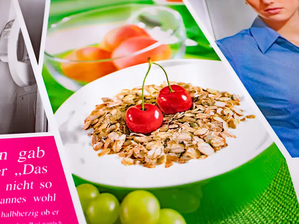 IMG_0133 A stack of printed brochures, produced using flexographic printing by DEBATIN DEBAPRINT, showcasing various designs. In the foreground, a bowl of muesli topped with two bright red cherries. The green background features gleaming peaches and grapes. Surrounding the central motif are additional images, including a partial face, a medical device, and a pink text block with white lettering.