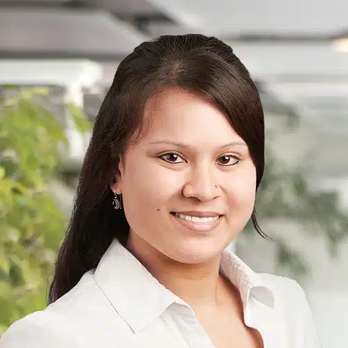 Close-up of Alexandra Böhmer, Purchasing Department at Debatin. A smiling woman with long dark hair and a nose piercing. She wears a white blouse, with blurred green plants and light structures in the background.