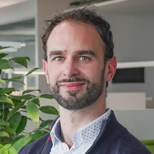 Portrait of Christophe Pille, a man with dark curly hair and a short beard. He wears a patterned light blue shirt and a dark pullover. Christophe Pille works in Sales Support at DEBATIN.