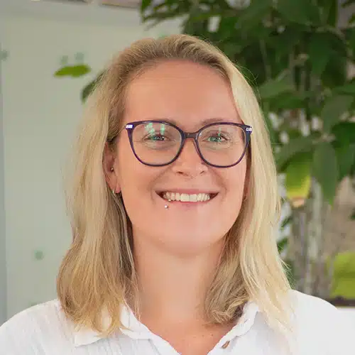 Portrait of Denise Raupp, Purchasing Department at Debatin. She smiles kindly, wearing glasses and a lip piercing. Her blonde hair is shoulder-length, and she wears a white top against a blurred green background.