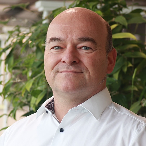 Portrait of Dr. Karsten Tauber. He is a middle-aged man with a bald head and a light shirt, smiling kindly at the camera. A green leafy plant is visible in the background.