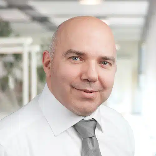 Head and shoulders portrait of Helmut Roth, Purchasing Department at Debatin. He looks directly at the camera with a slight smile. Bald man wearing a white shirt and a grey tie. The blurred background suggests an office setting.