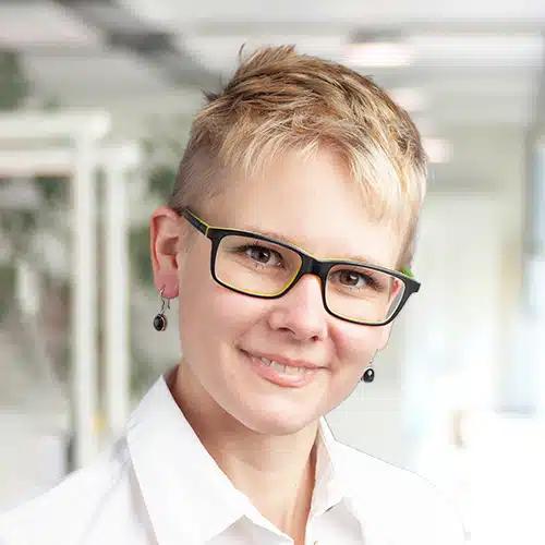 Melanie Kost, Shipping Department at Debatin, smiles warmly at the camera. Her short platinum blonde hair and stylish glasses with neon-green accents give her an individual touch.