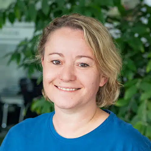 Close-up of Michaela Breßer, working as Sales Consultant / Customer Service at Debatin. A smiling woman with short blonde hair wearing a blue T-shirt. Blurry green plants are visible in the background.