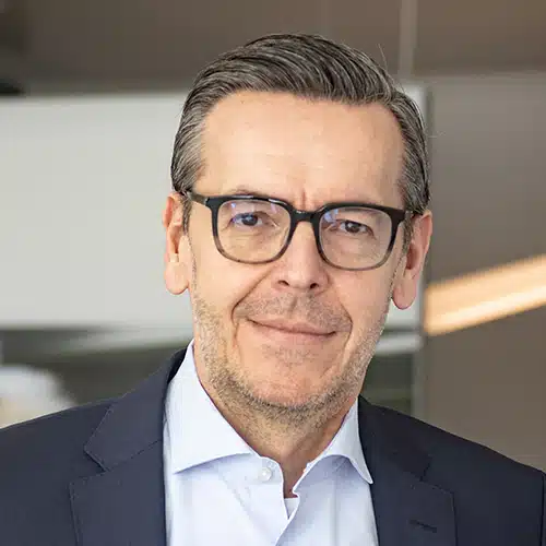 Portrait of Oliver Kosalla, Head of Purchasing at Debatin. He wears modern glasses, a dark blue jacket over a light blue shirt, and looks directly into the camera. His face shows a neat beard and greying hair.