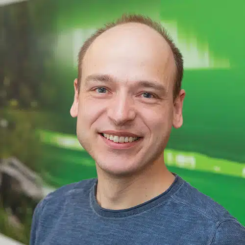 Portrait of Oliver Tröber, Development Team at Debatin. He smiles kindly at the camera, wears a simple blue shirt and has short light hair. The background shows a blurred green surface.