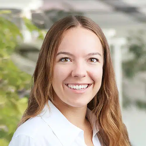 Close-up of Patricia Polzer, working as Sales Consultant / Customer Service at Debatin. A friendly smiling woman with long brown hair wearing a white blouse. The blurred background shows bright and green tones, suggesting an office environment.