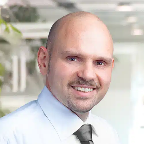 Head and shoulders portrait of Thomas Harböck, Purchasing Department at Debatin. He smiles at the camera, bald with a neat moustache. He wears a light shirt and a dark tie, with blurred green accents in the background.