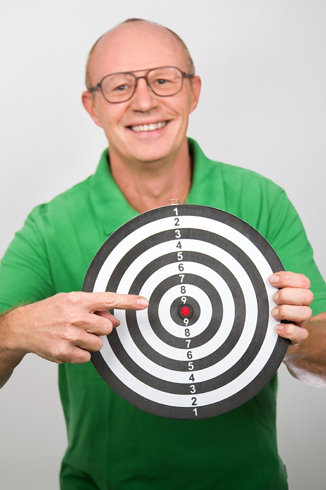 DEBATIN_Produktsparten_Headerbild_DEBADEINS_Gerald Frank_Zielscheibe Gerald Frank is wearing a green shirt and holding up a colorful dartboard.