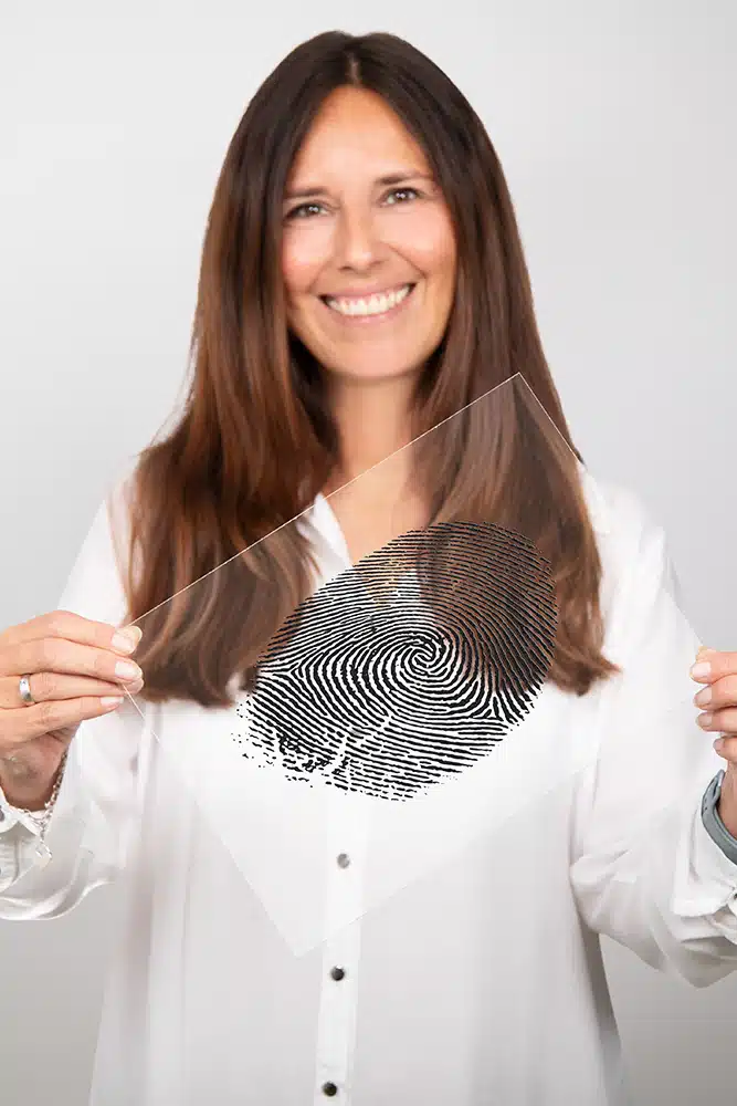 DEBATIN_Produktsparten_Headerbild_DEBADEINS_Hillenbrand_Fingerabdruck Gaby Hillenbrand is wearing a white blouse and holding up a fingerprint engraved into a plexiglass panel.