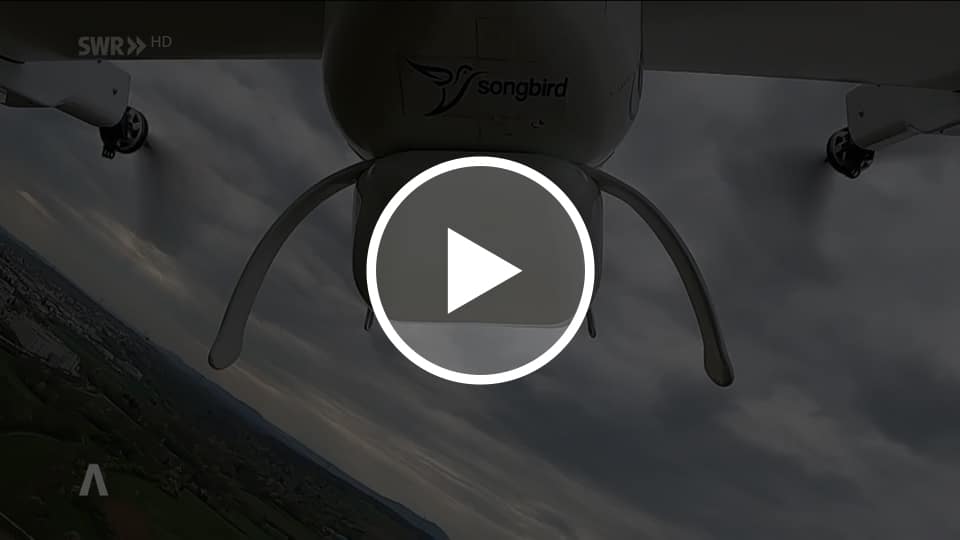 The underside of a small drone is seen from below during flight. A camera pod and landing gear are visible. The logo and text “songbird” are printed on the aircraft body. Below, a landscape with fields and buildings appears tilted, under a cloudy sky.