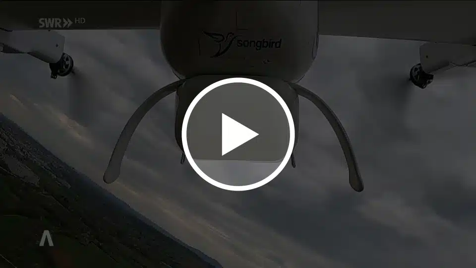 The underside of a small drone is seen from below during flight. A camera pod and landing gear are visible. The logo and text “songbird” are printed on the aircraft body. Below, a landscape with fields and buildings appears tilted, under a cloudy sky.