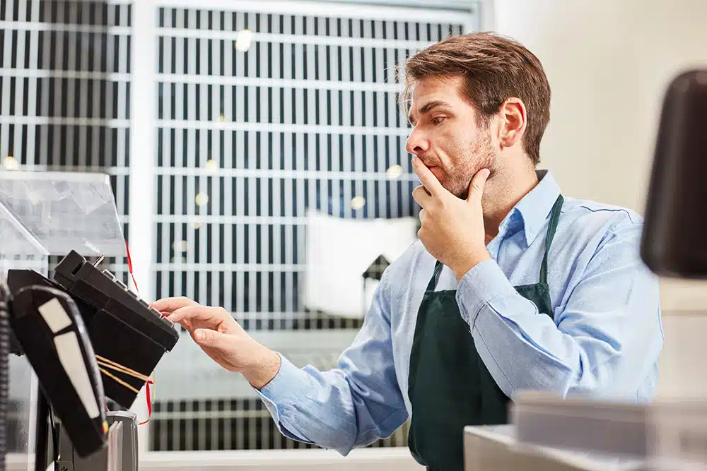 A cashier standing thoughtfully at a till, touching their chin with the left hand while the other rests on the keypad.