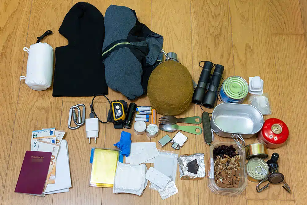 Close-up of travel and emergency equipment spread out on a wooden floor, including cash, a passport, a water bottle, a headlamp, batteries, canned food, and a first-aid kit.