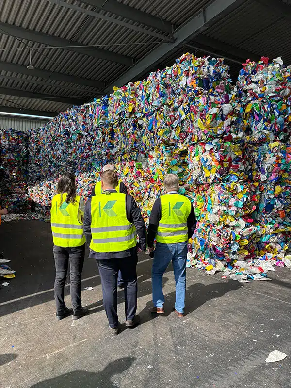 Four people in hi-vis vests, seen from behind, are looking at a huge wall of compressed, multicoloured plastic waste in a warehouse.