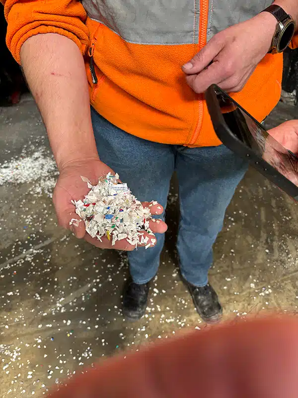 A person in an orange work jacket is holding a handful of shredded plastic scraps. Many small plastic pieces are also scattered on the floor.