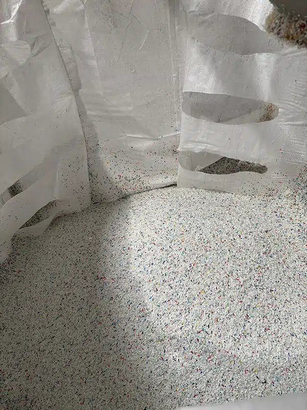A large white sack is filled with tiny, colourful plastic fragments. The different colours of the fragments indicate recycled material.