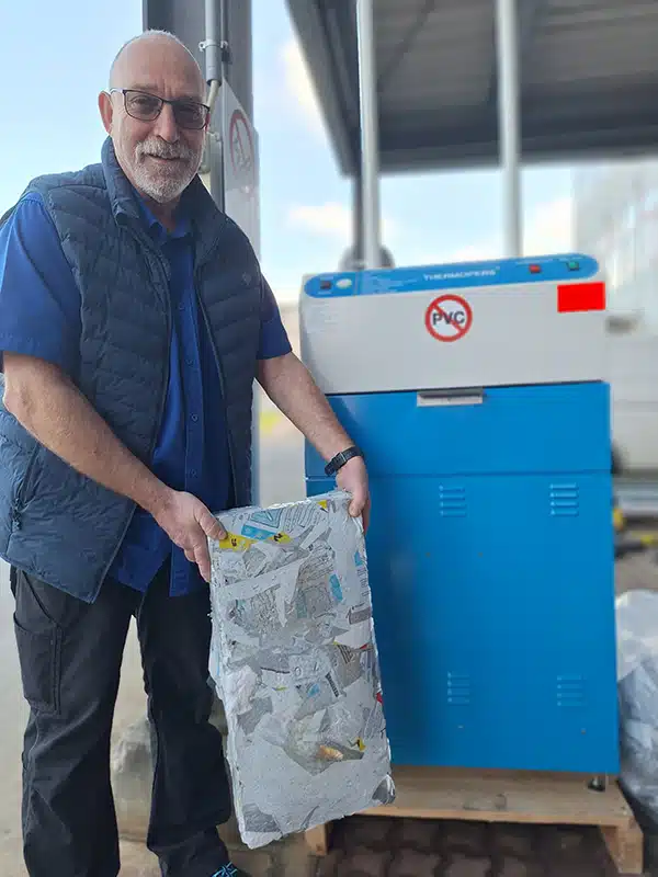 debatin_press_bioscientia Jürgen Heß, Head of Supply Chain Management at Bioscientia Healthcare GmbH, stands next to the blue DEBATIN plastic recycling baler. He is holding a compacted bale of recycled plastic material in his hands. In the background, a covered outdoor area of an industrial building can be seen.